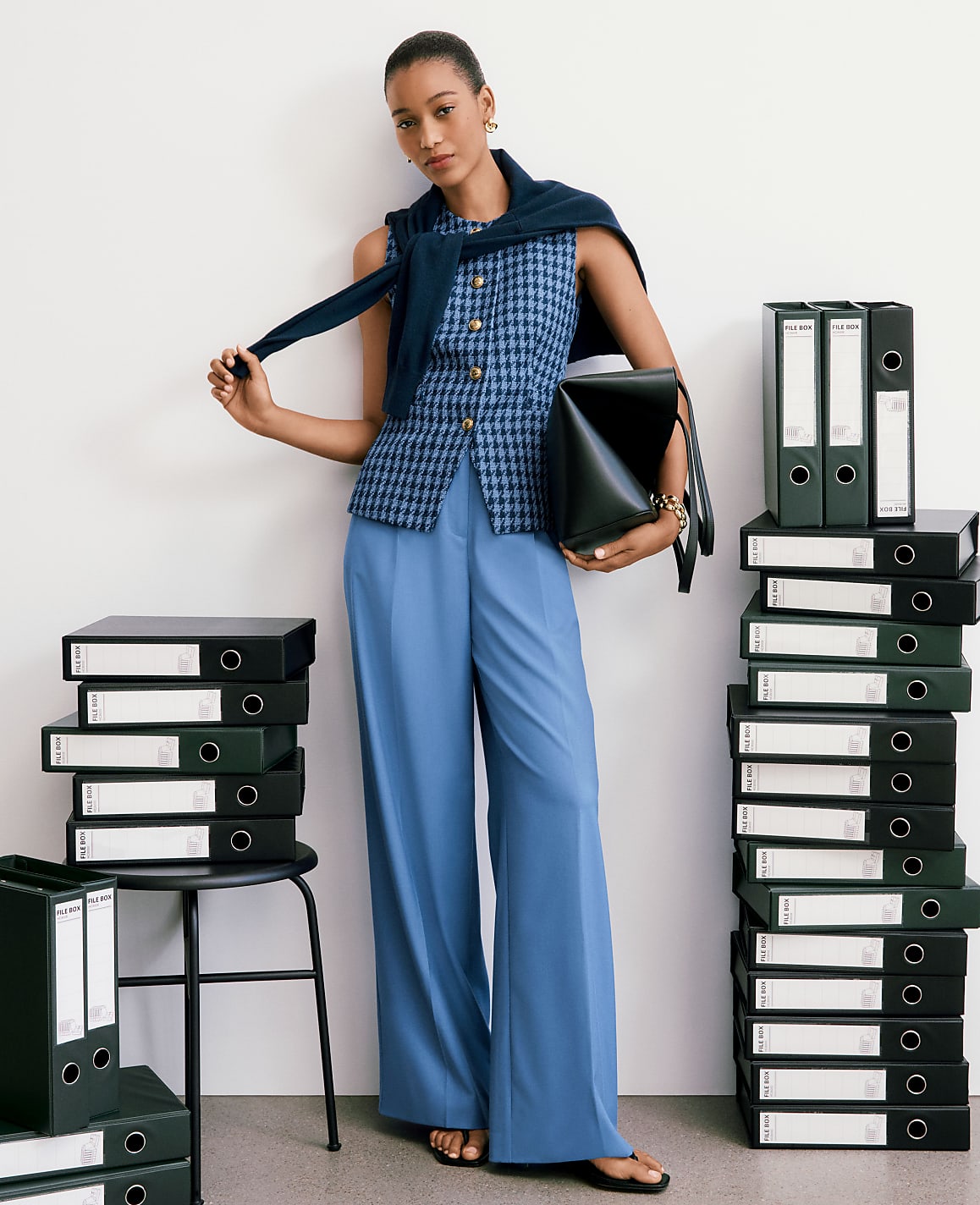 A woman in blue pants and a patterned vest stands between stacks of black binders against a white wall.