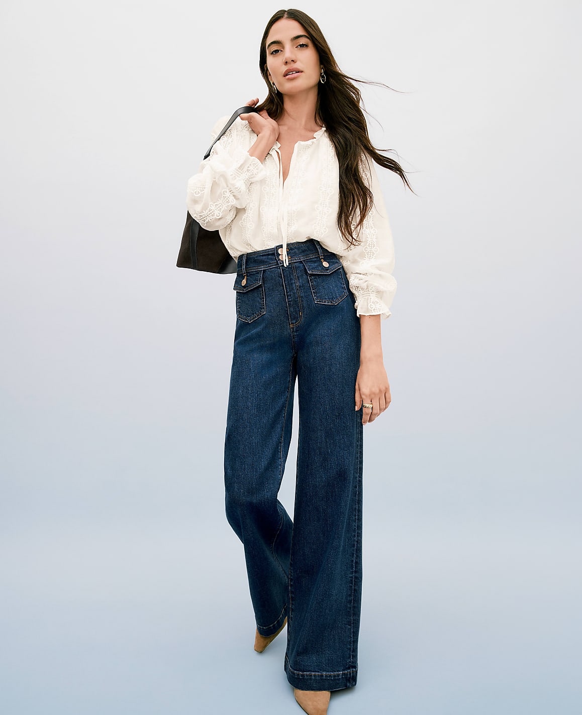 A woman in a white blouse and wide-leg jeans stands against a light blue background, with a dark jacket draped over her shoulder.