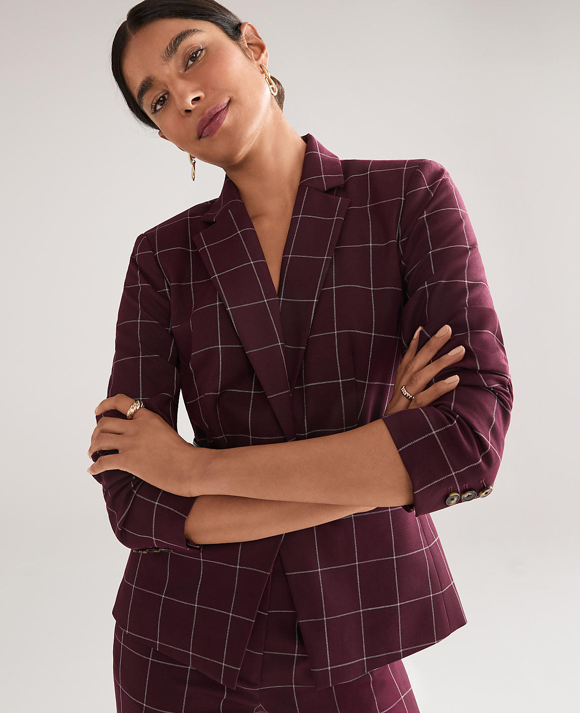 A woman in a burgundy plaid suit stands with her arms crossed against a plain gray background.