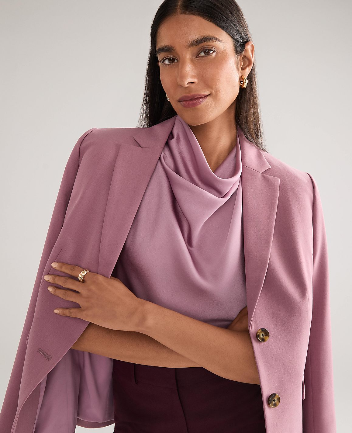 A woman in a mauve blazer and cowl-neck top poses against a plain, light gray background.