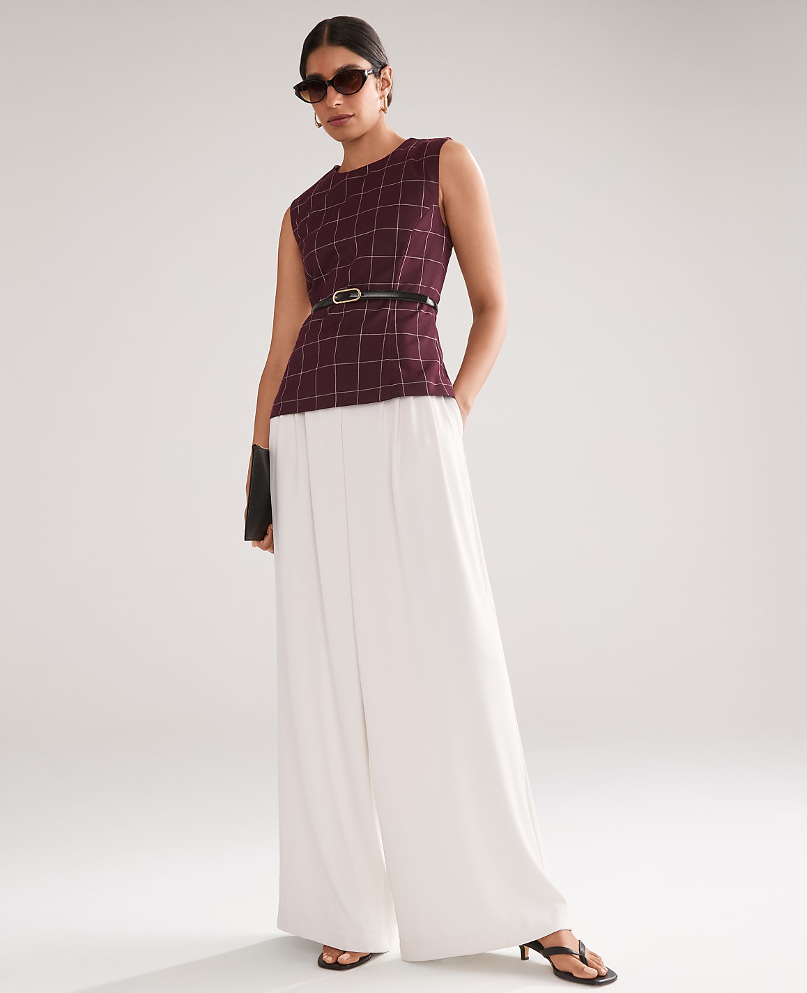 A woman in a maroon plaid top and white wide-leg pants stands against a plain, light gray background.