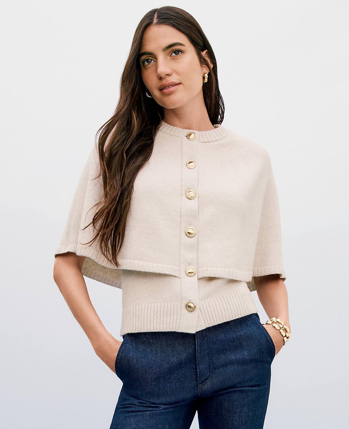 A woman in a cream-colored capelet and sweater stands against a plain, light background.