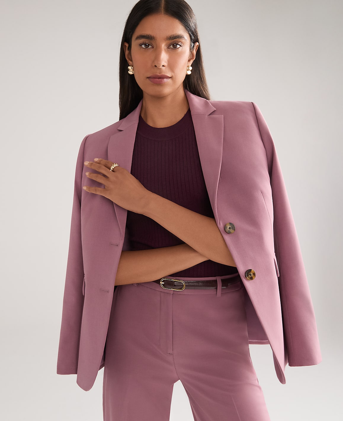 A woman in a mauve blazer and matching pants stands against a plain, light gray background.