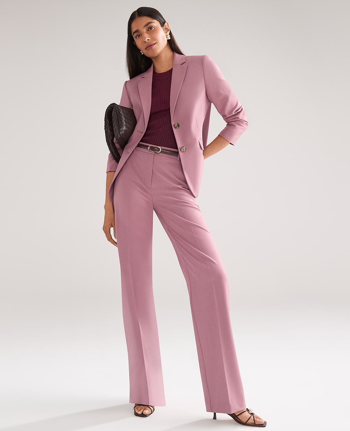 A woman in a mauve pantsuit and burgundy top stands with a clutch in her hand against a plain, light gray background.