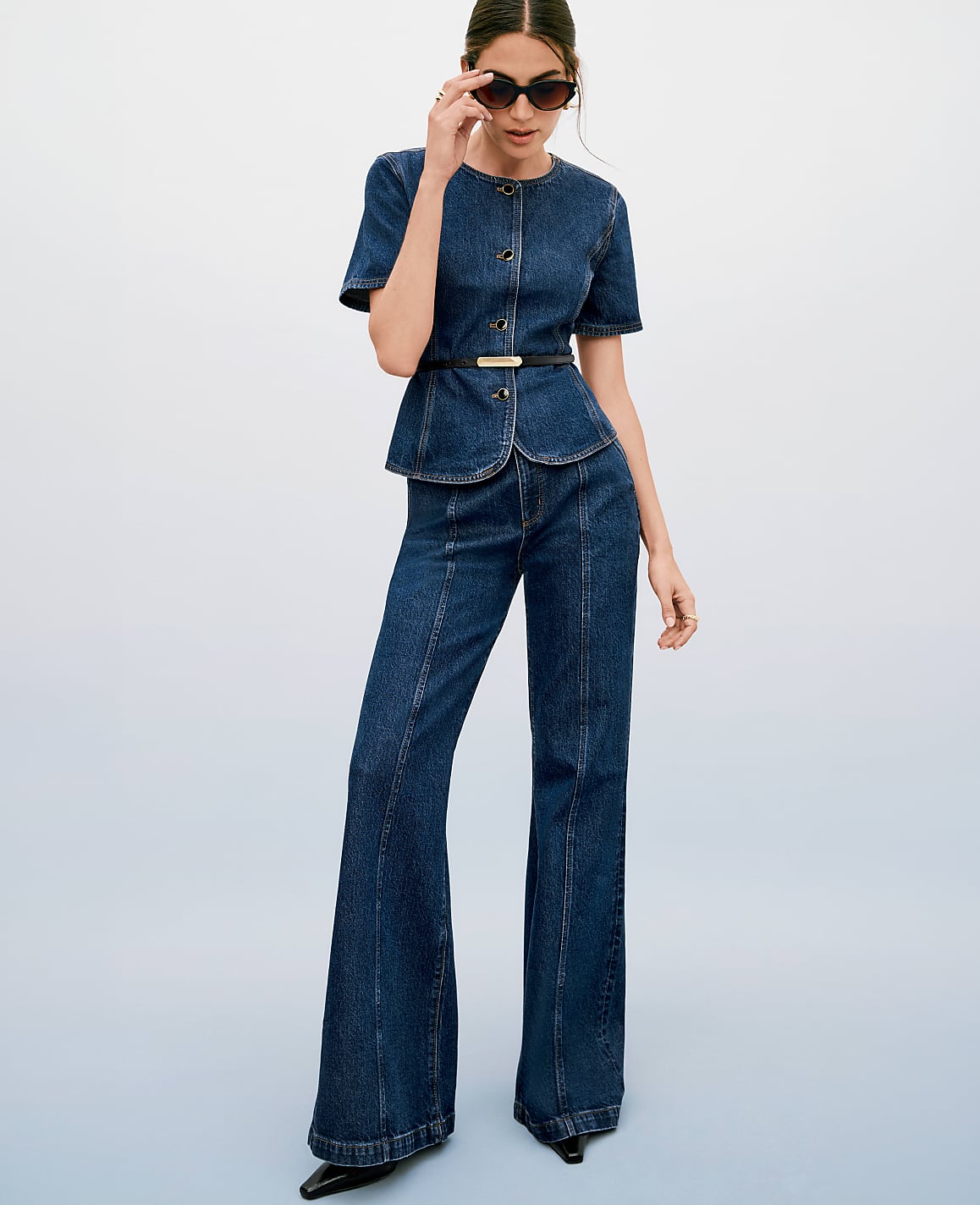 A woman in a denim peplum top and flared jeans poses against a gradient blue background.