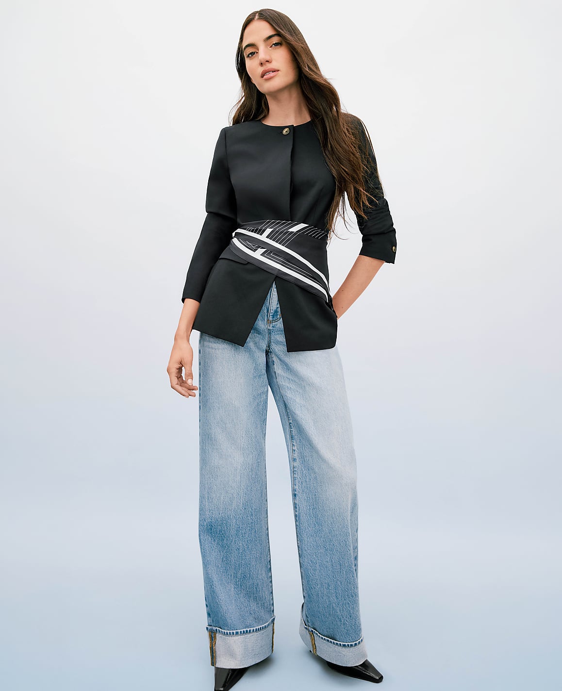 A woman in a black blazer and wide-leg jeans stands against a soft blue gradient background.