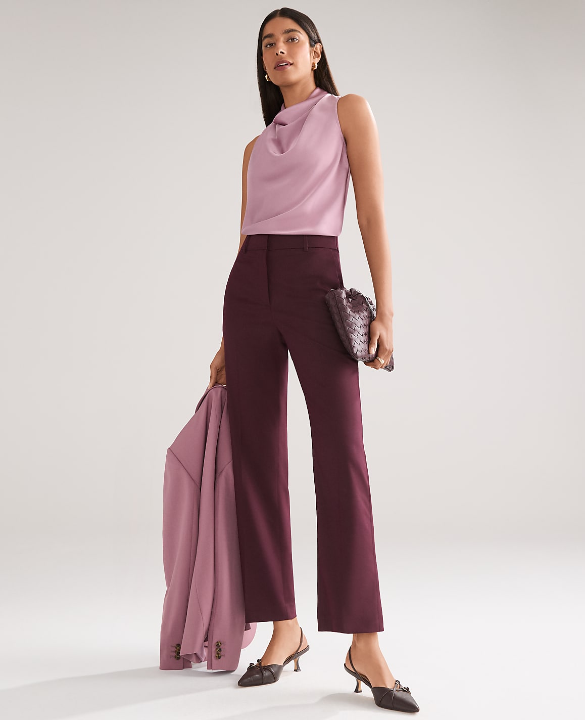 A woman in a lilac cowl-neck top and burgundy wide-leg pants stands against a plain, light gray background, holding a matching blazer and a patterned clutch.