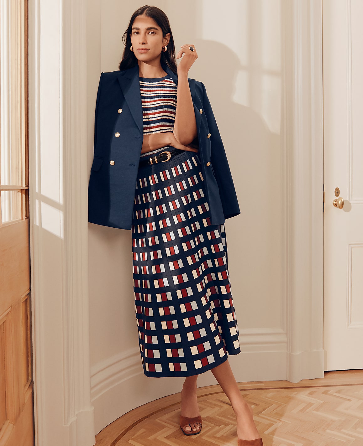 A woman in a striped top, navy blazer, and patterned skirt stands in a hallway with a wooden floor and white doors.