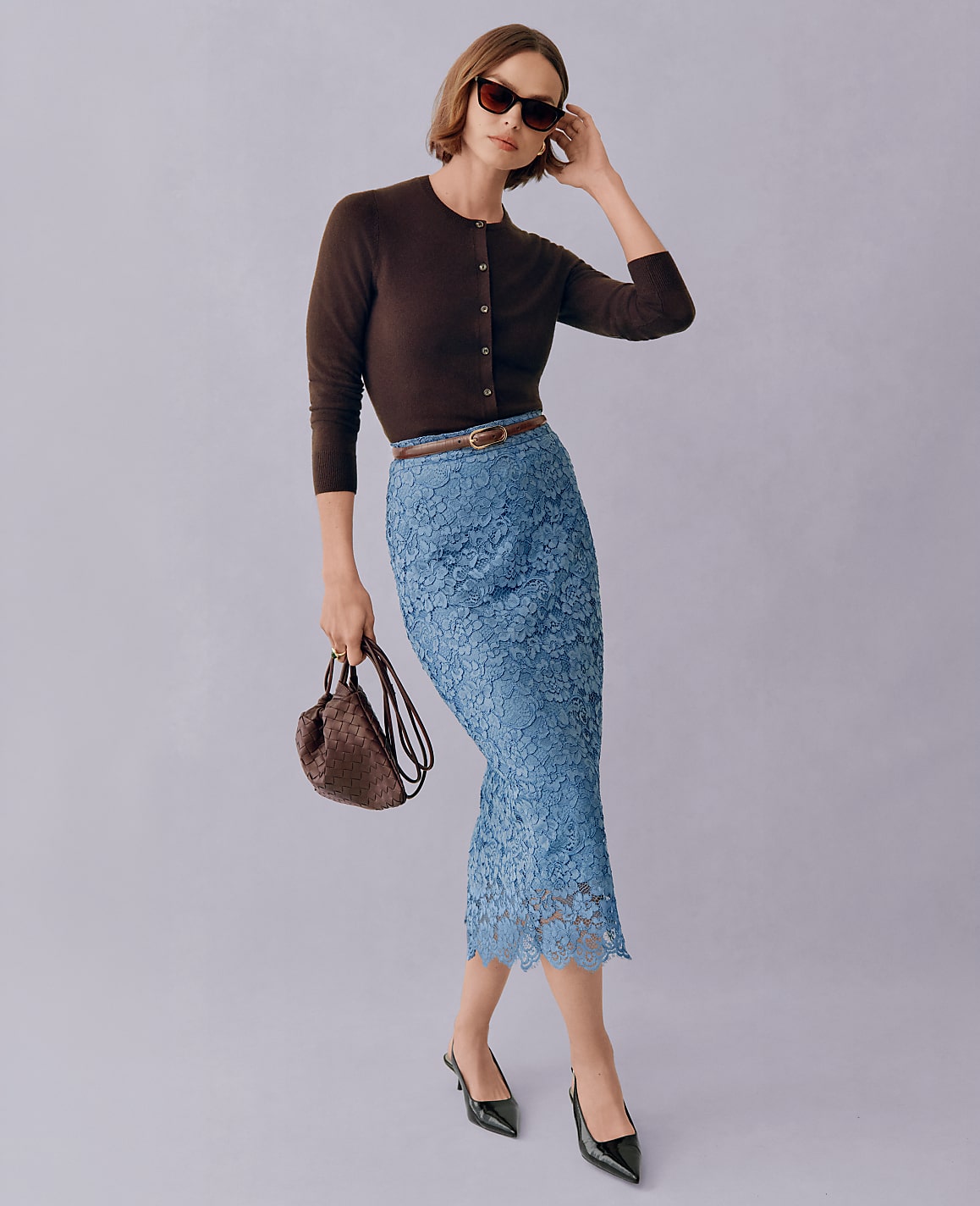 A woman in a brown cardigan and blue lace skirt poses with a handbag against a plain background.