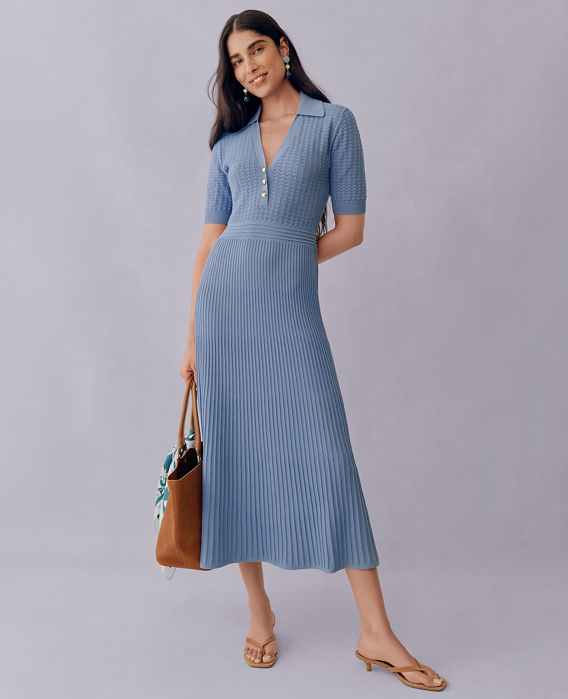 A woman in a blue knit dress and sandals stands with a tote bag against a plain purple background.