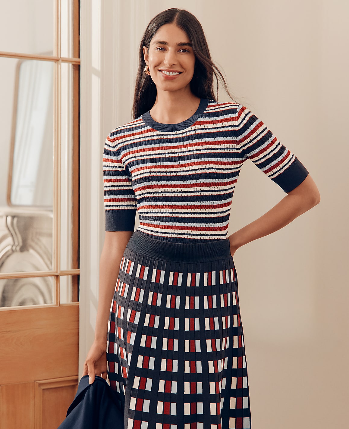 A smiling woman in a striped top and patterned skirt stands in a well-lit room with a doorway to her left.