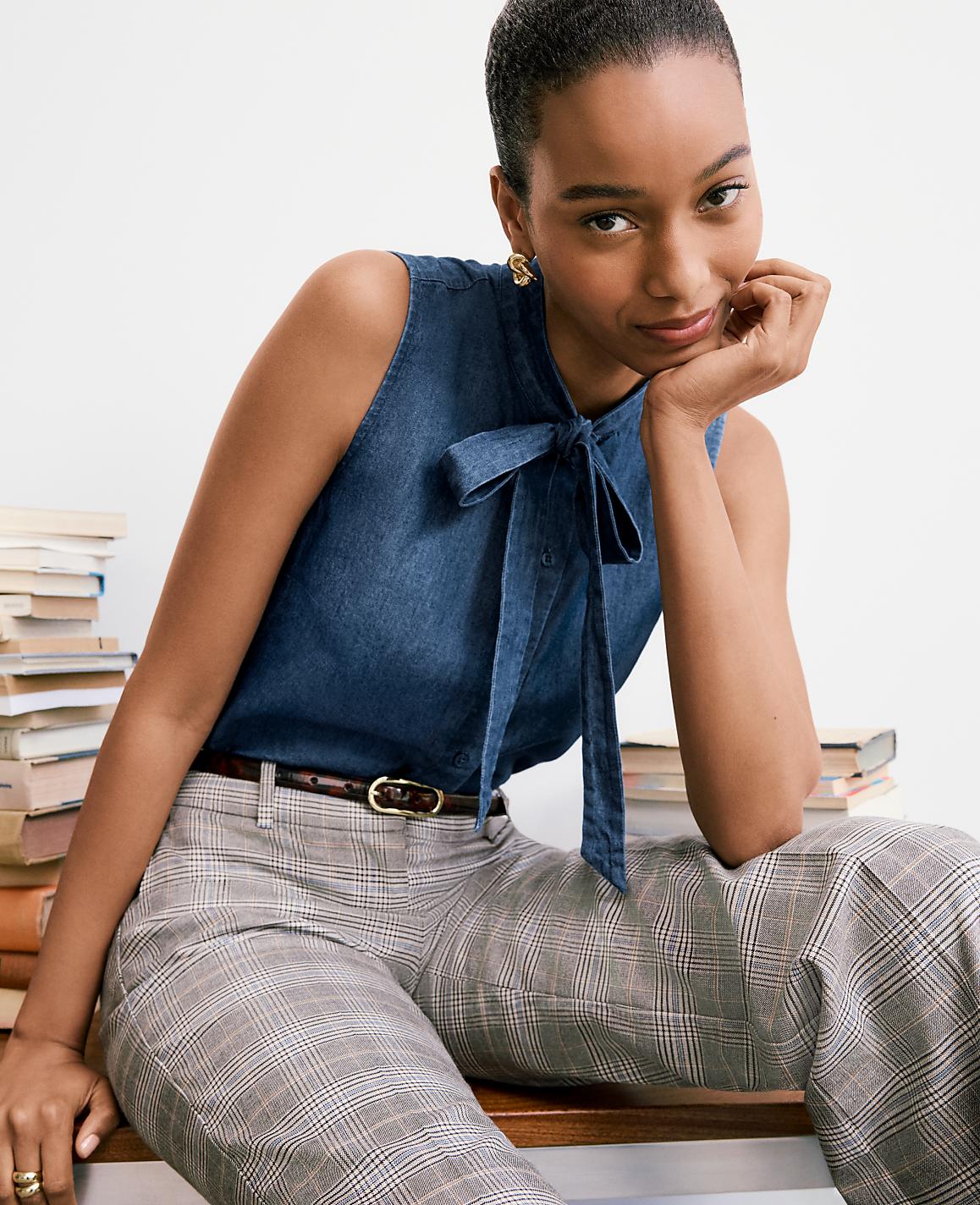 A woman in a denim bow-neck top and plaid pants rests her chin on her hand, with a stack of books behind her.