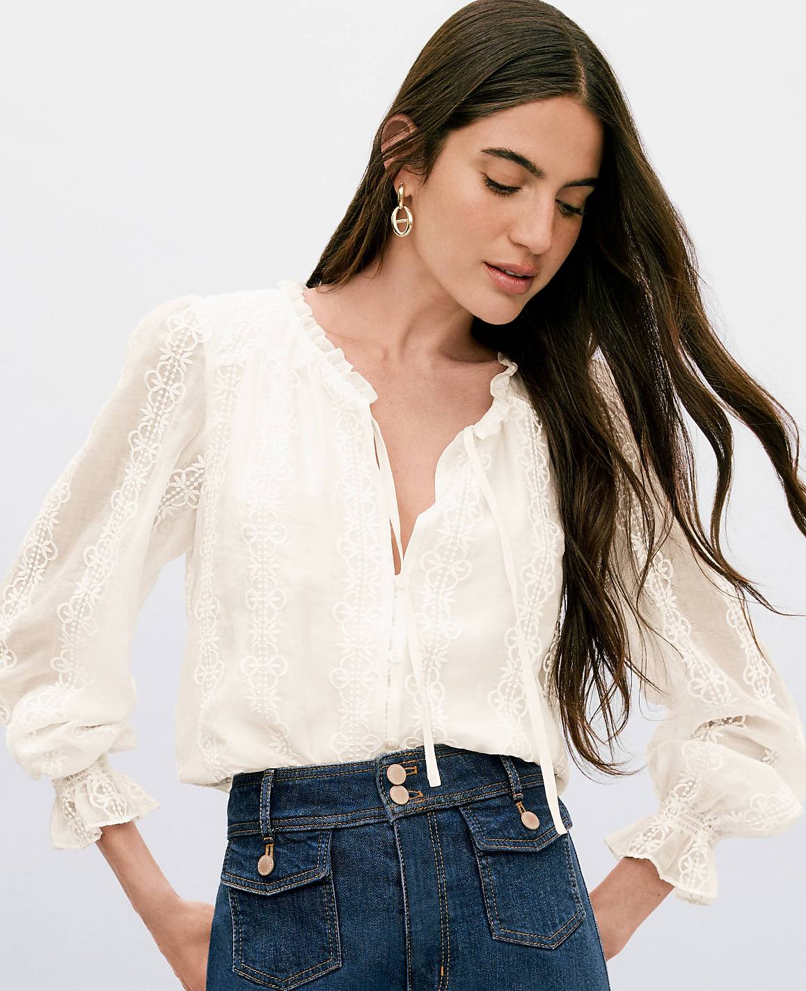 A woman with long dark hair wears a white lace-trimmed blouse and dark denim jeans against a plain white background.