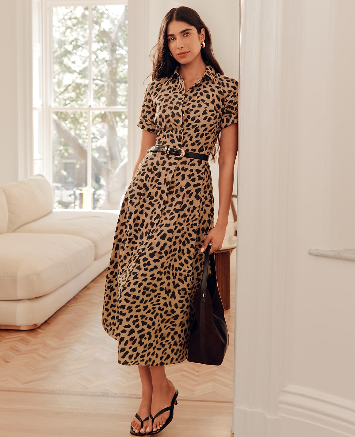 A woman in a leopard print dress stands in a bright room with a sofa in the background.