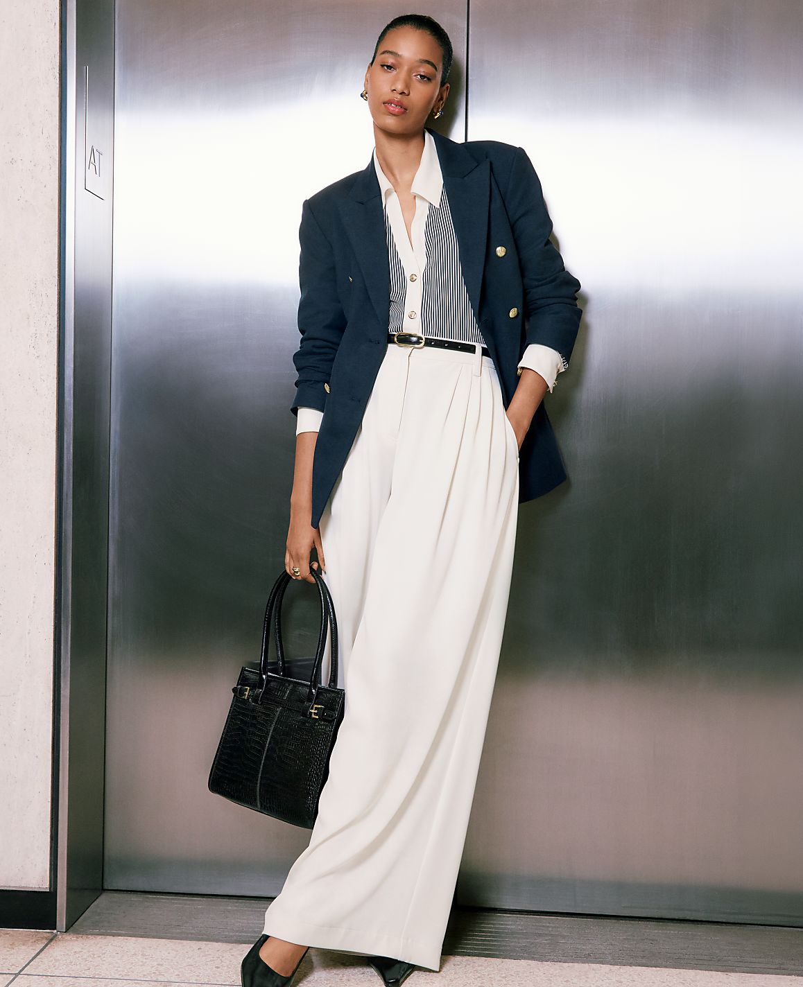 A woman in a navy blazer and white pants stands in front of a brushed metal elevator door, holding a black handbag.