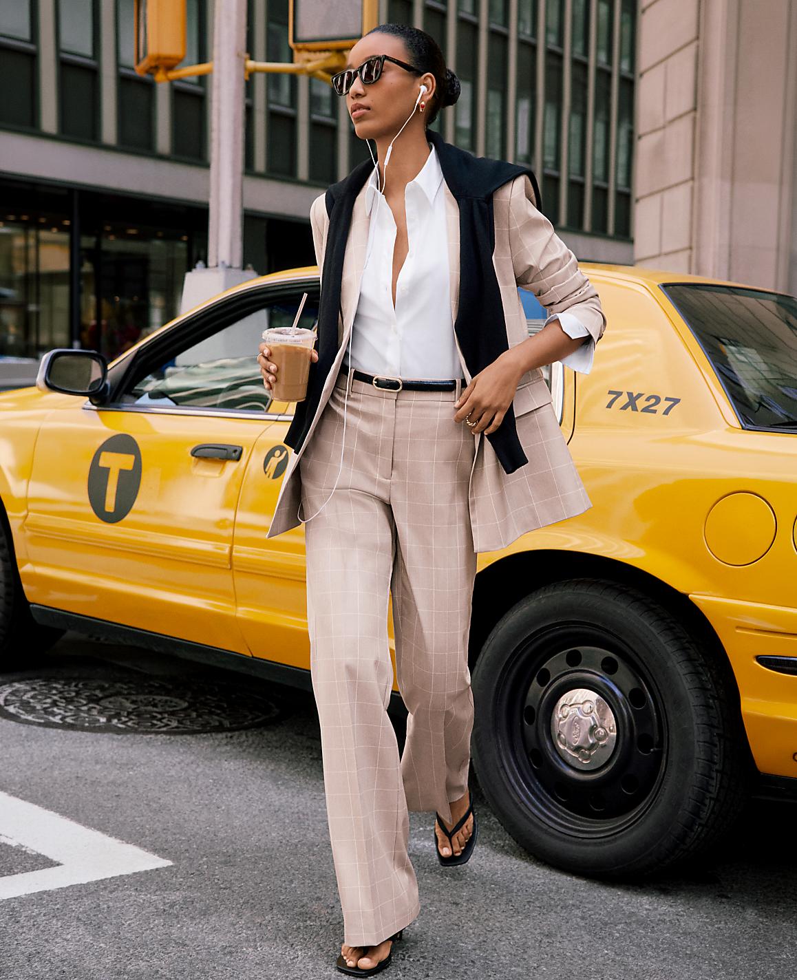 A woman in a tan plaid suit walks past a yellow taxi in a city street.