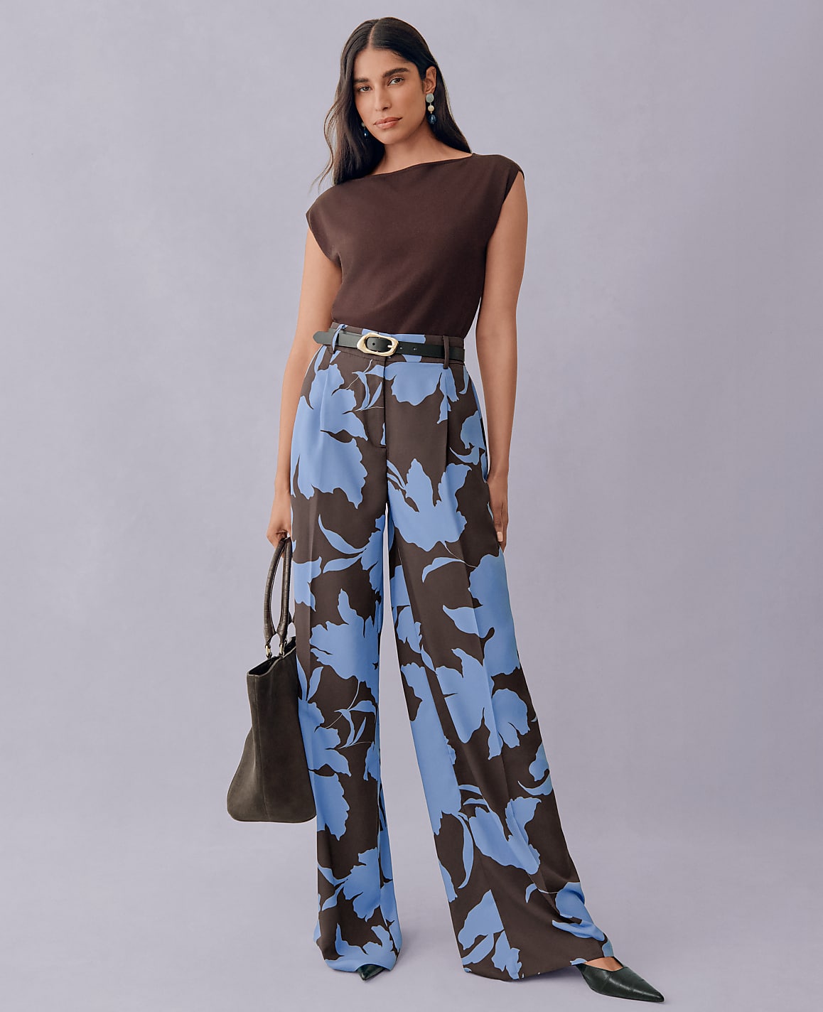 A woman in a brown top and floral wide-leg pants stands against a plain purple background, holding a dark handbag.