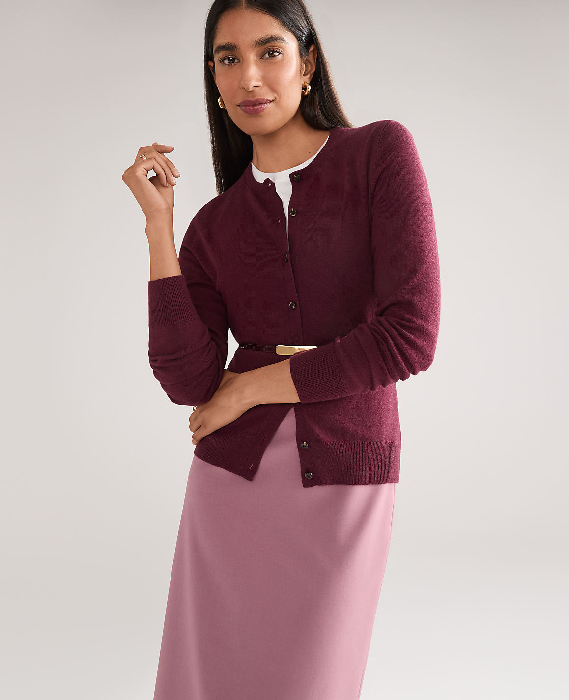 A woman in a burgundy cardigan and pink skirt stands against a plain gray background.