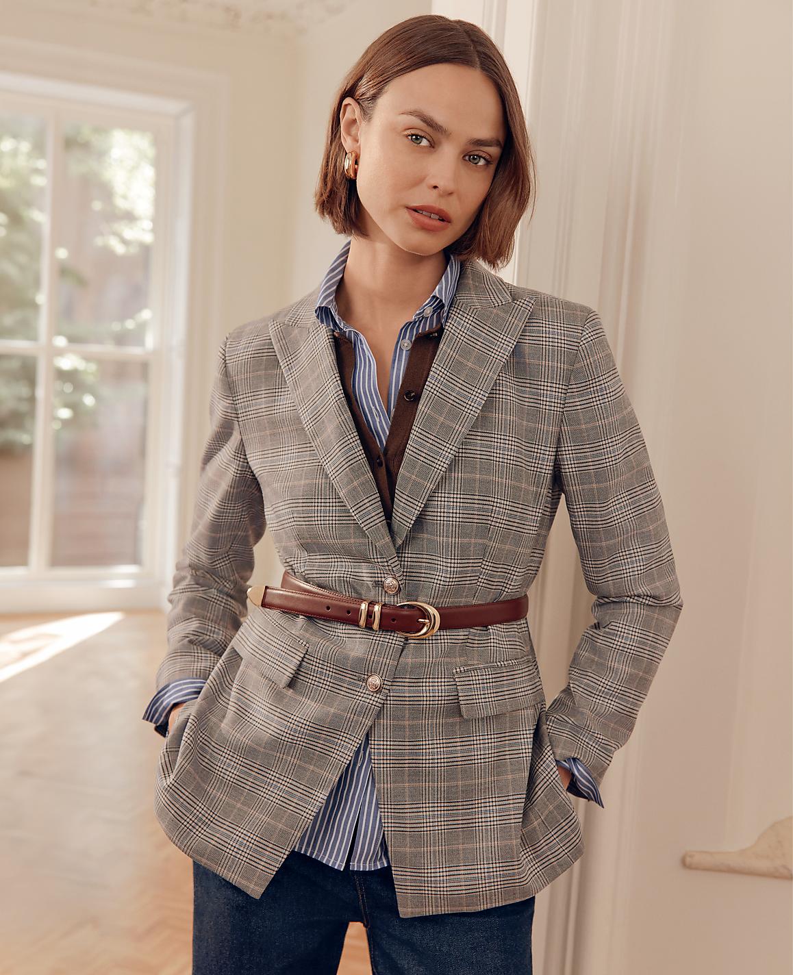 A woman in a plaid blazer and striped shirt stands in a well-lit room with a window in the background.