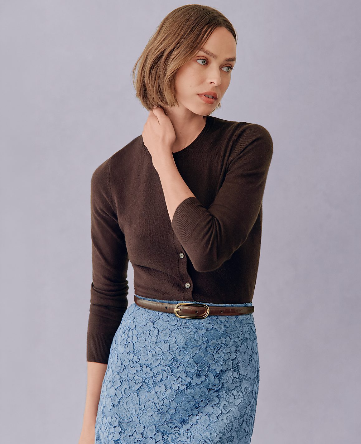 A woman in a brown cardigan and blue lace skirt stands against a plain purple background.