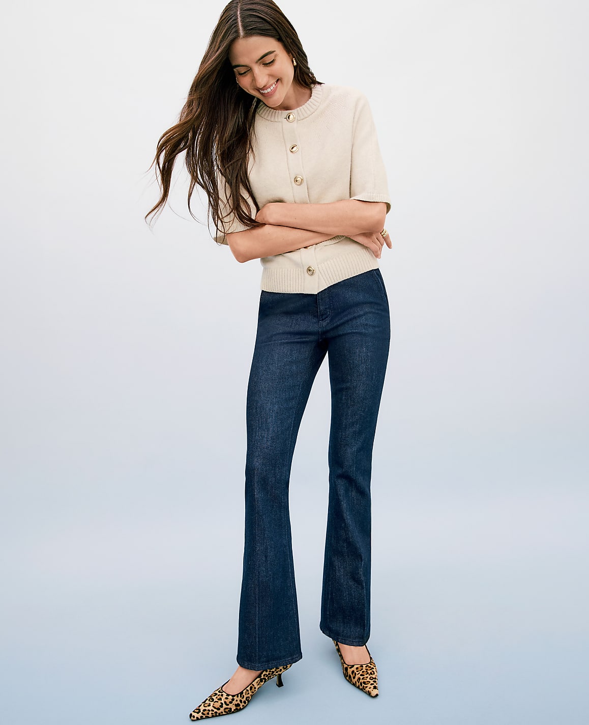 A smiling woman in a cream cardigan and dark flare jeans stands against a soft blue background.