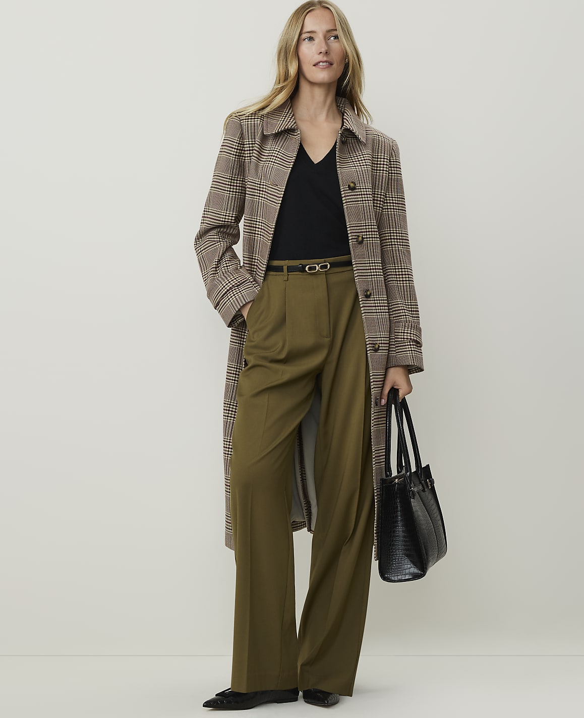 A woman in a plaid coat and olive green pants stands against a plain, light-colored background, holding a black handbag.
