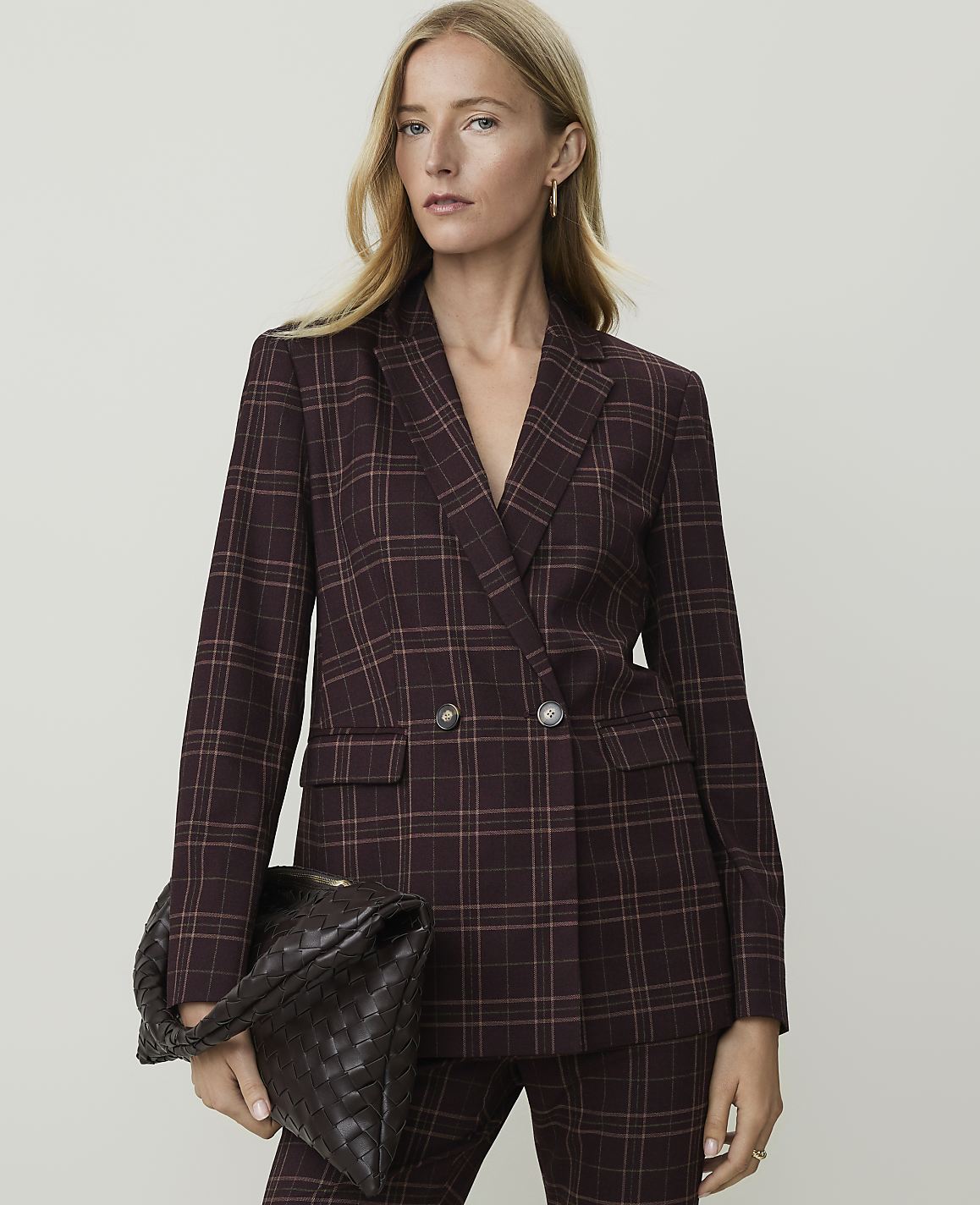 A woman in a burgundy plaid blazer and matching pants holds a woven clutch in front of a plain gray background.