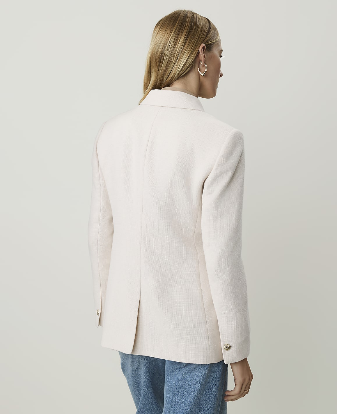A woman in a light pink blazer and blue jeans stands against a plain, light-colored background.
