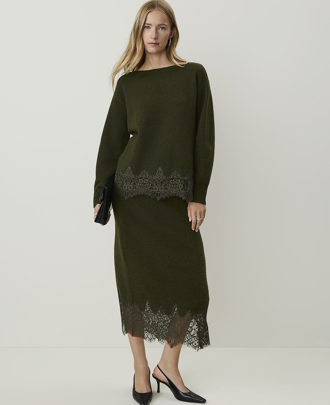 A woman in a green knit sweater and skirt with lace trim stands against a plain white background, holding a black clutch.