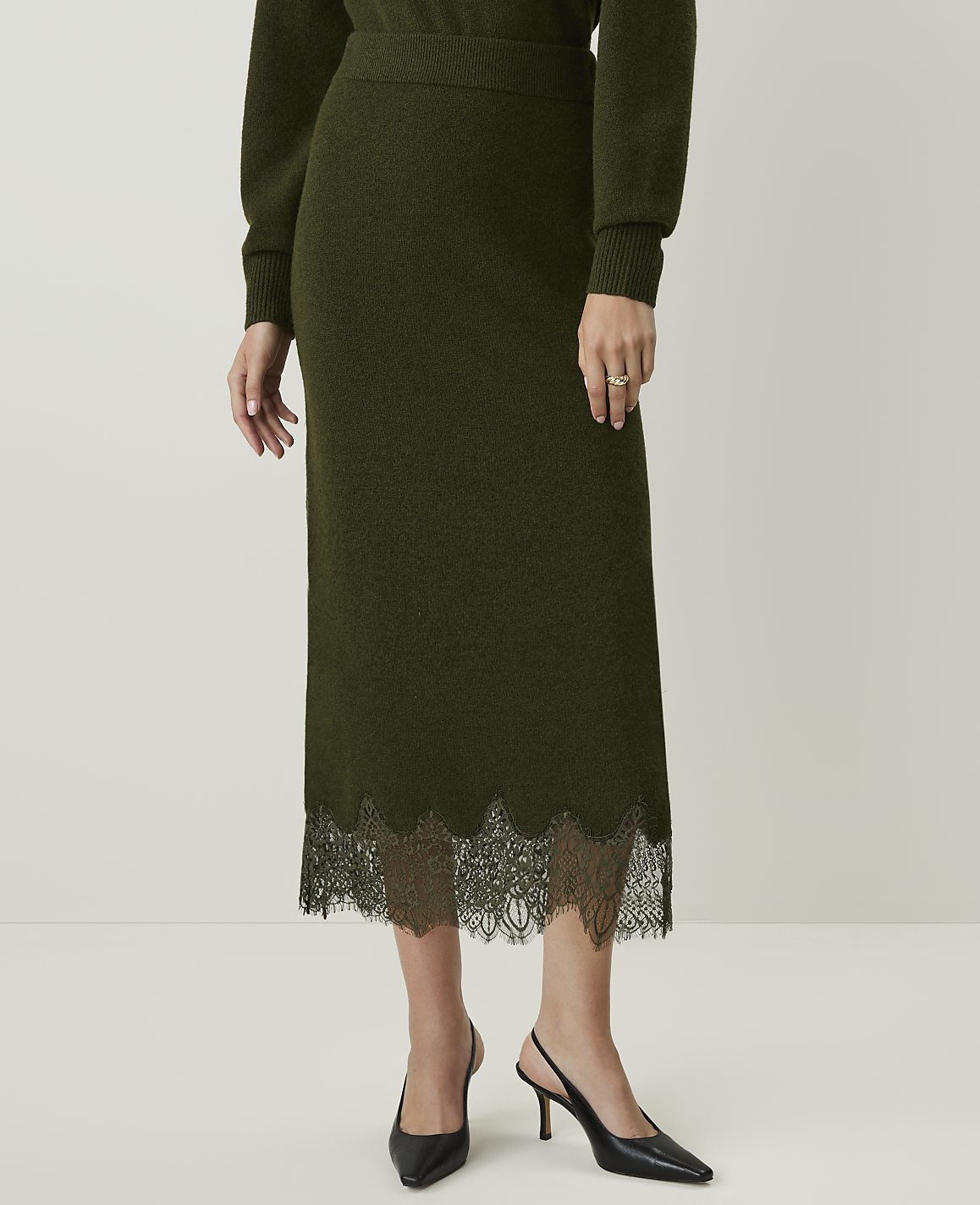 A person stands in a plain white studio, wearing a dark green knit midi skirt with a black lace hem and black slingback heels.