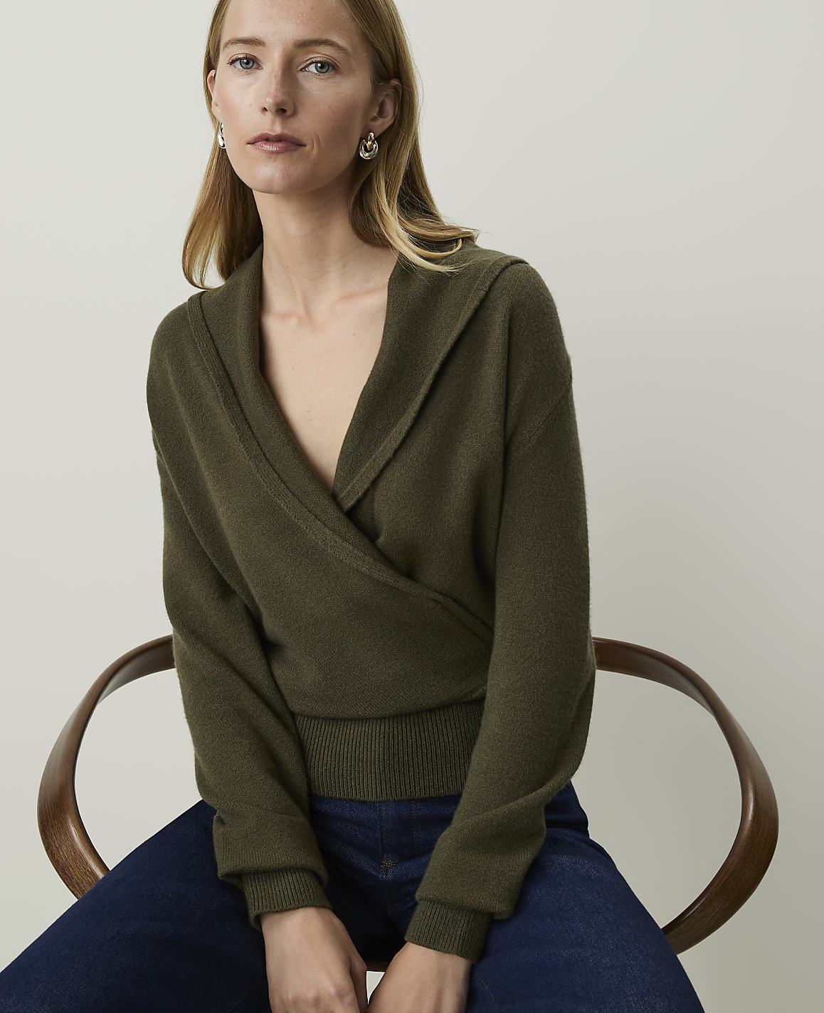 A woman in a green wrap sweater and blue jeans sits on a wooden chair against a plain background.