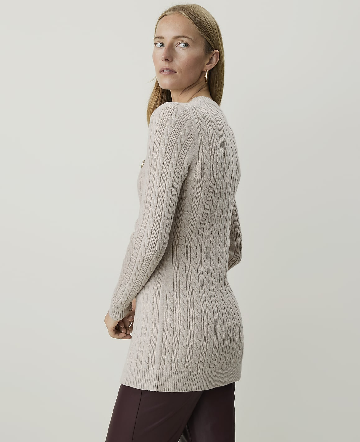 A woman in a beige cable-knit sweater and burgundy pants stands against a plain, light gray background.