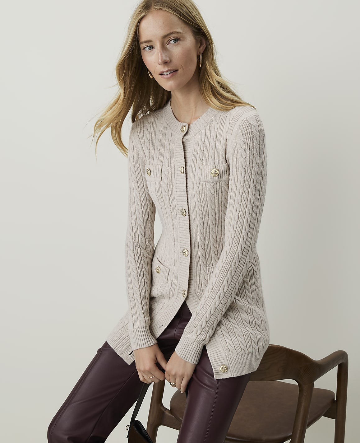 A woman in a cream cable-knit cardigan and burgundy pants sits on a wooden chair against a plain, light background.