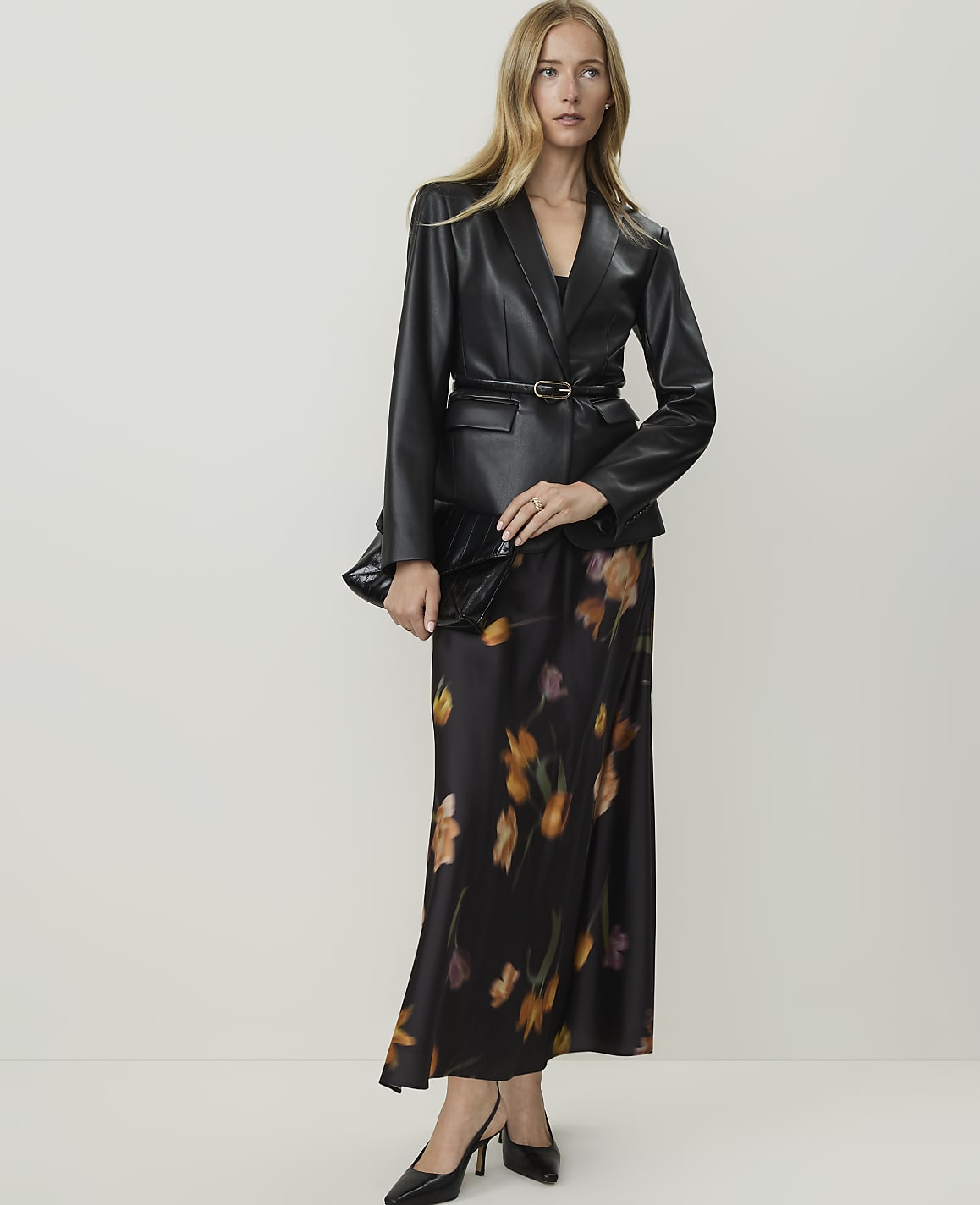 A woman in a black leather blazer and a floral print skirt stands against a plain white background.
