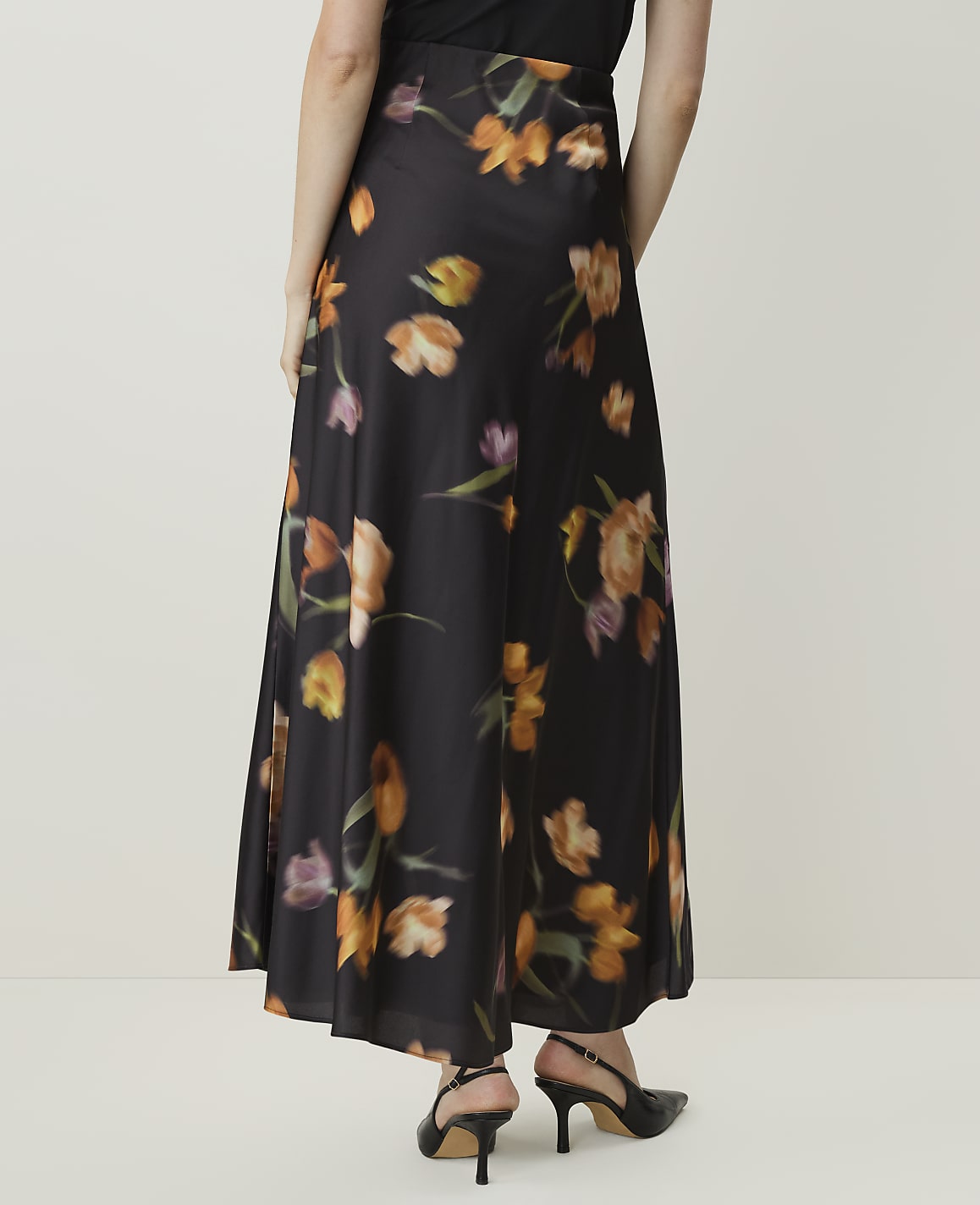 A person wearing a long, black floral skirt with orange and purple flowers stands against a plain, light-colored background.
