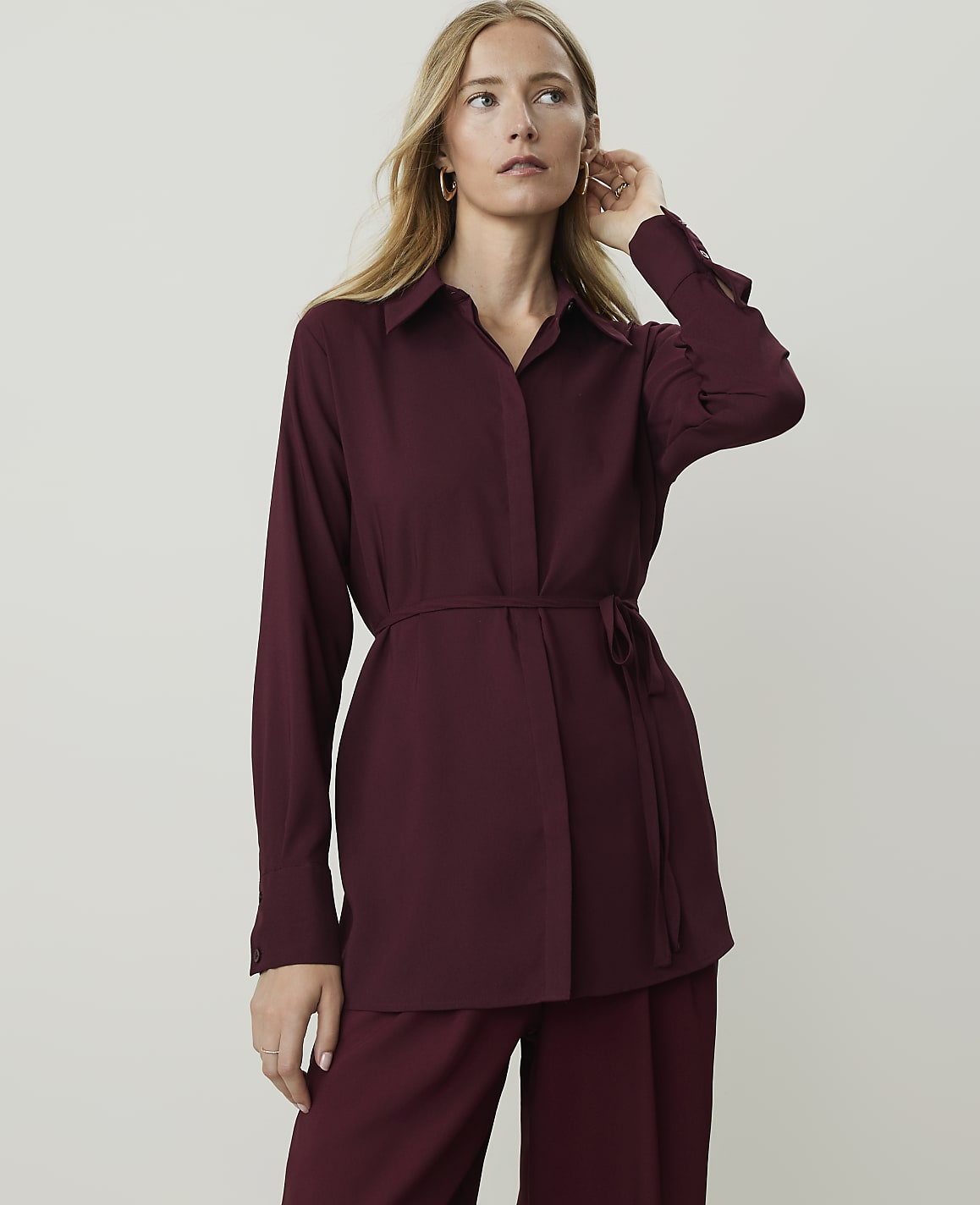 A woman in a burgundy tunic and pants stands against a plain, light gray background.