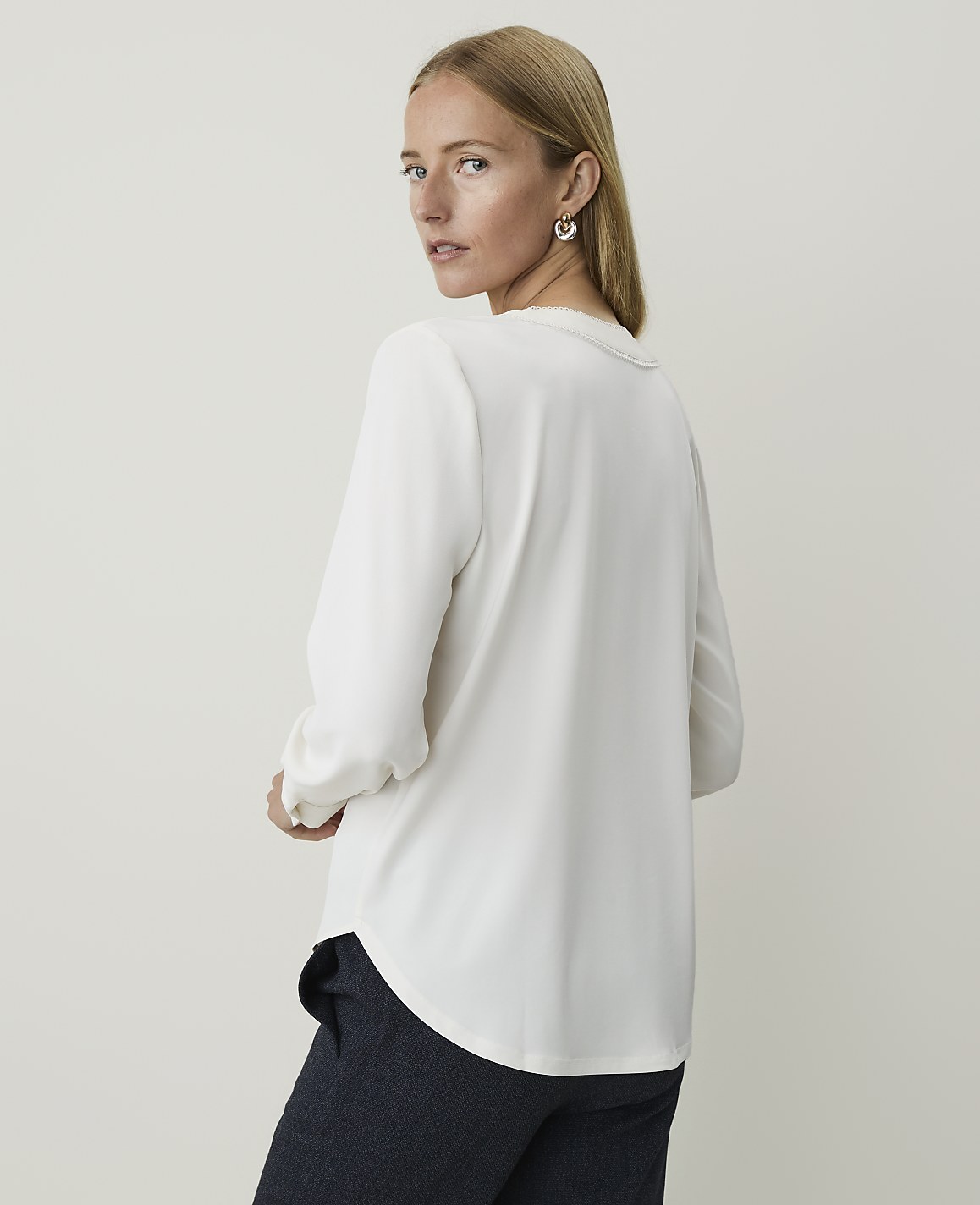 A woman in a white blouse and dark pants stands against a plain, light background.