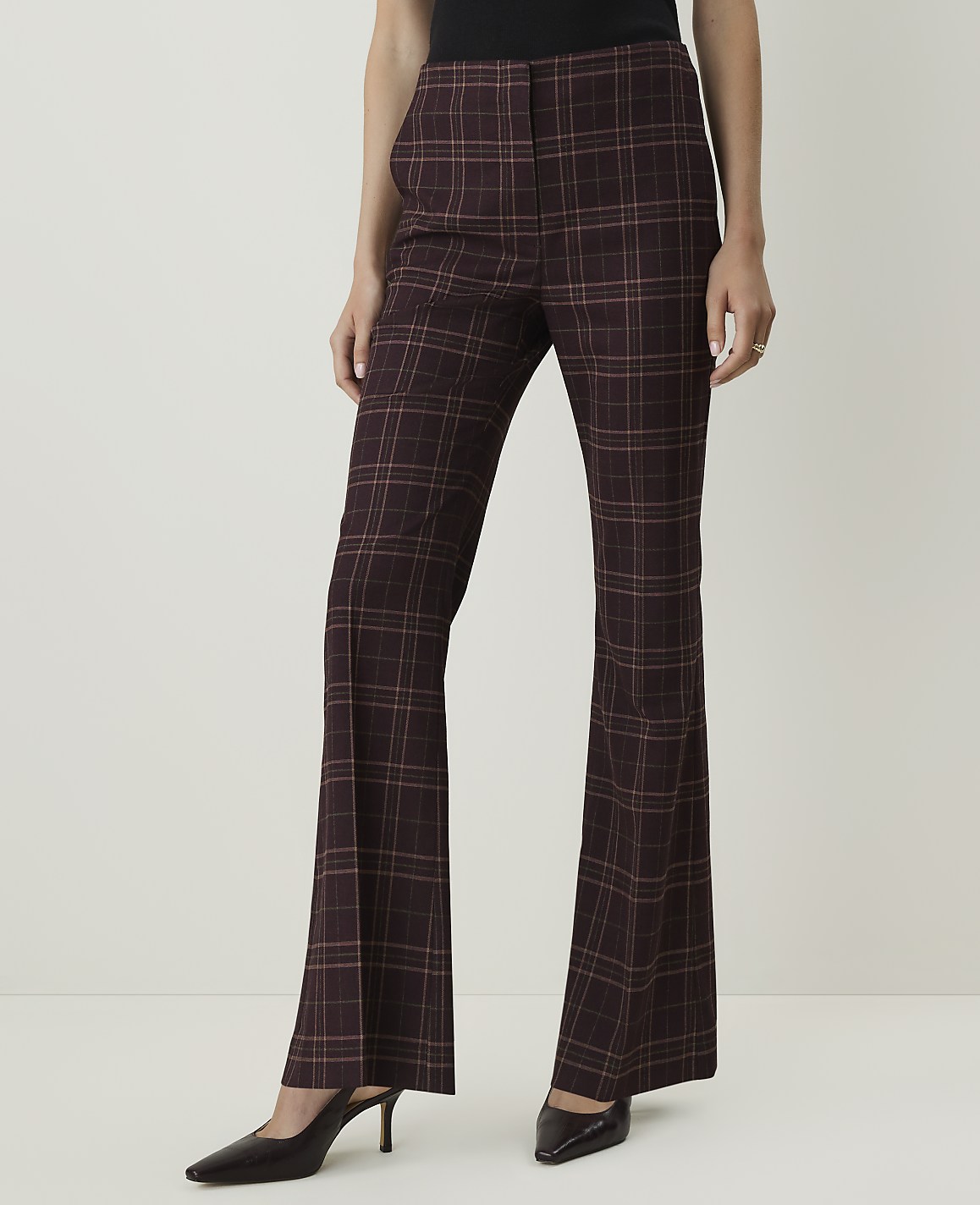 A person wearing maroon plaid flared pants and dark pointed-toe heels stands against a plain white background.