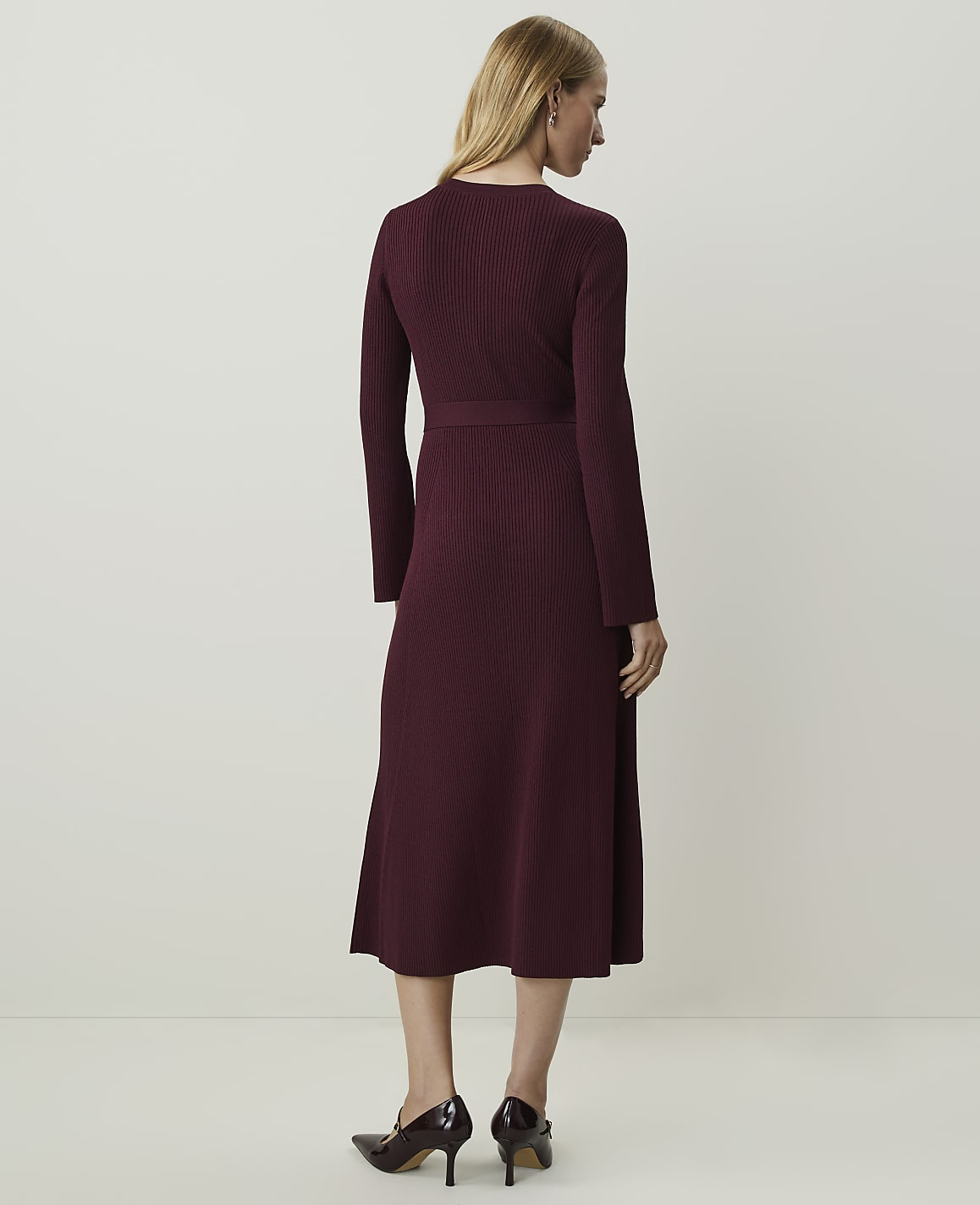 A woman in a burgundy ribbed midi dress and heels stands against a plain, light-colored background.