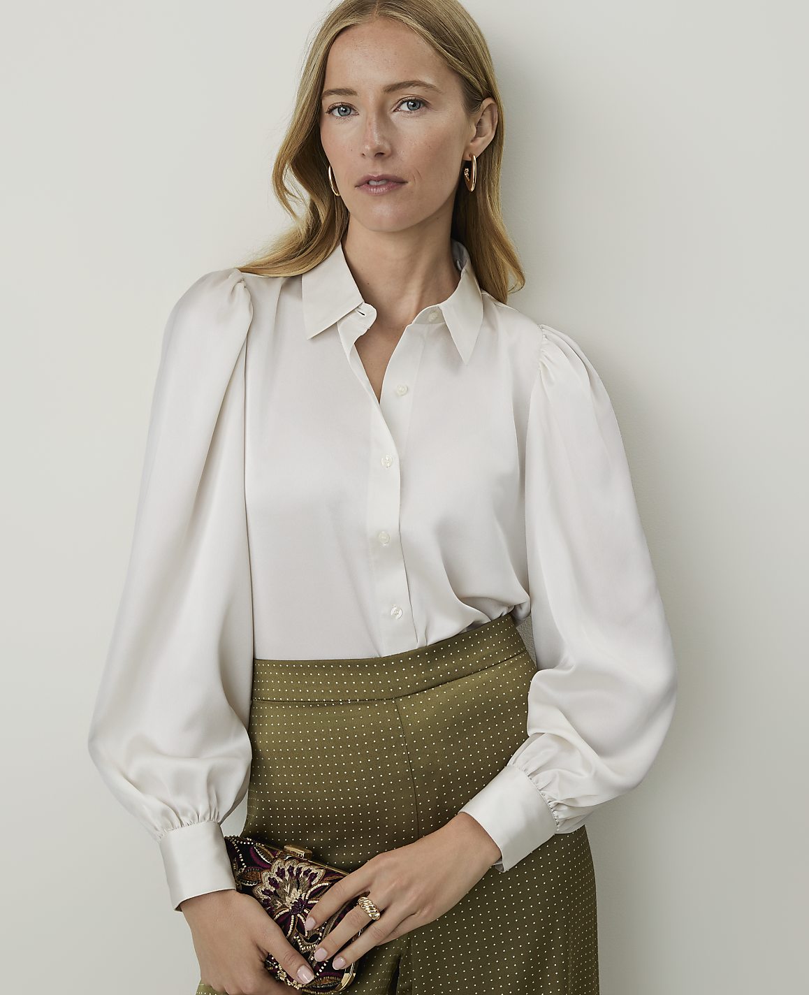 A woman in a white blouse and patterned pants holds a clutch in front of a plain background.