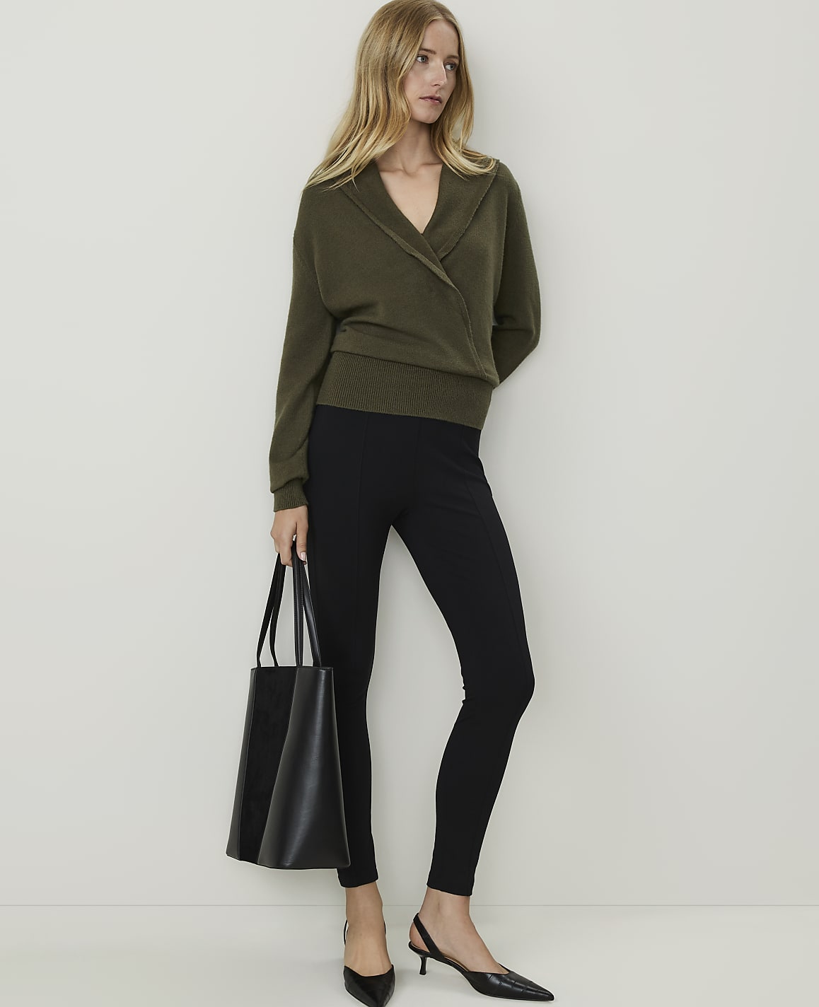 A woman in a green wrap sweater and black leggings stands with a black tote bag against a plain white background.