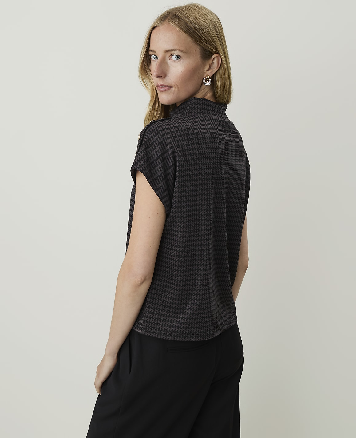 A woman with blonde hair and fair skin, wearing a dark patterned top and black pants, is shown from the back against a plain, light gray background.