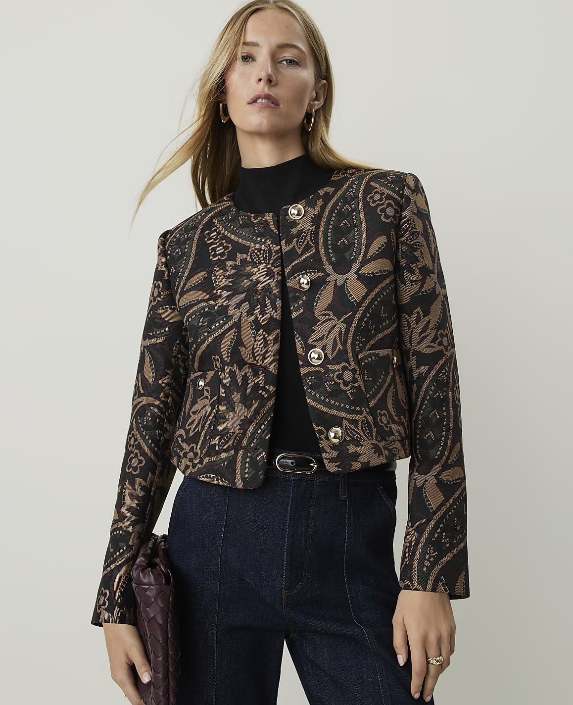A woman in a patterned jacket and dark jeans stands against a plain, light background.