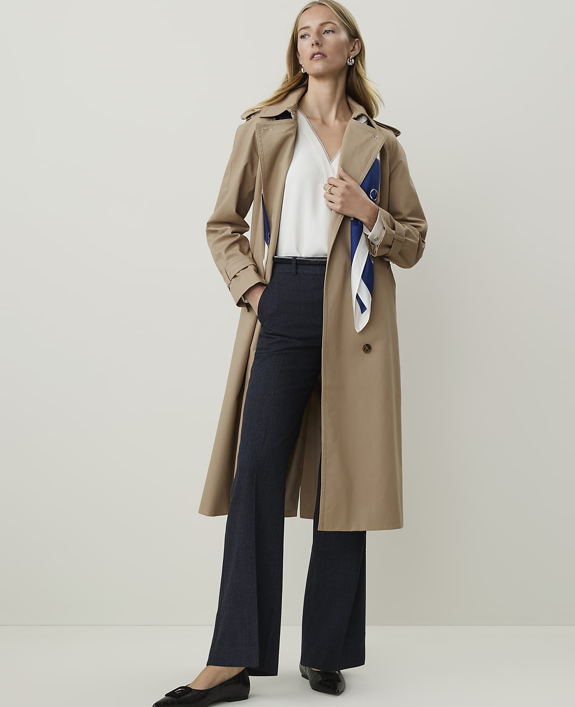 A woman in a tan trench coat and dark pinstripe pants stands against a plain, light gray background.