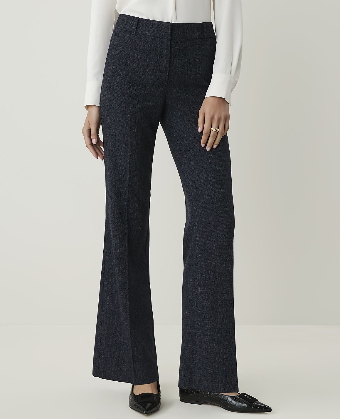 A person in a white blouse and dark patterned pants stands against a plain, light background.