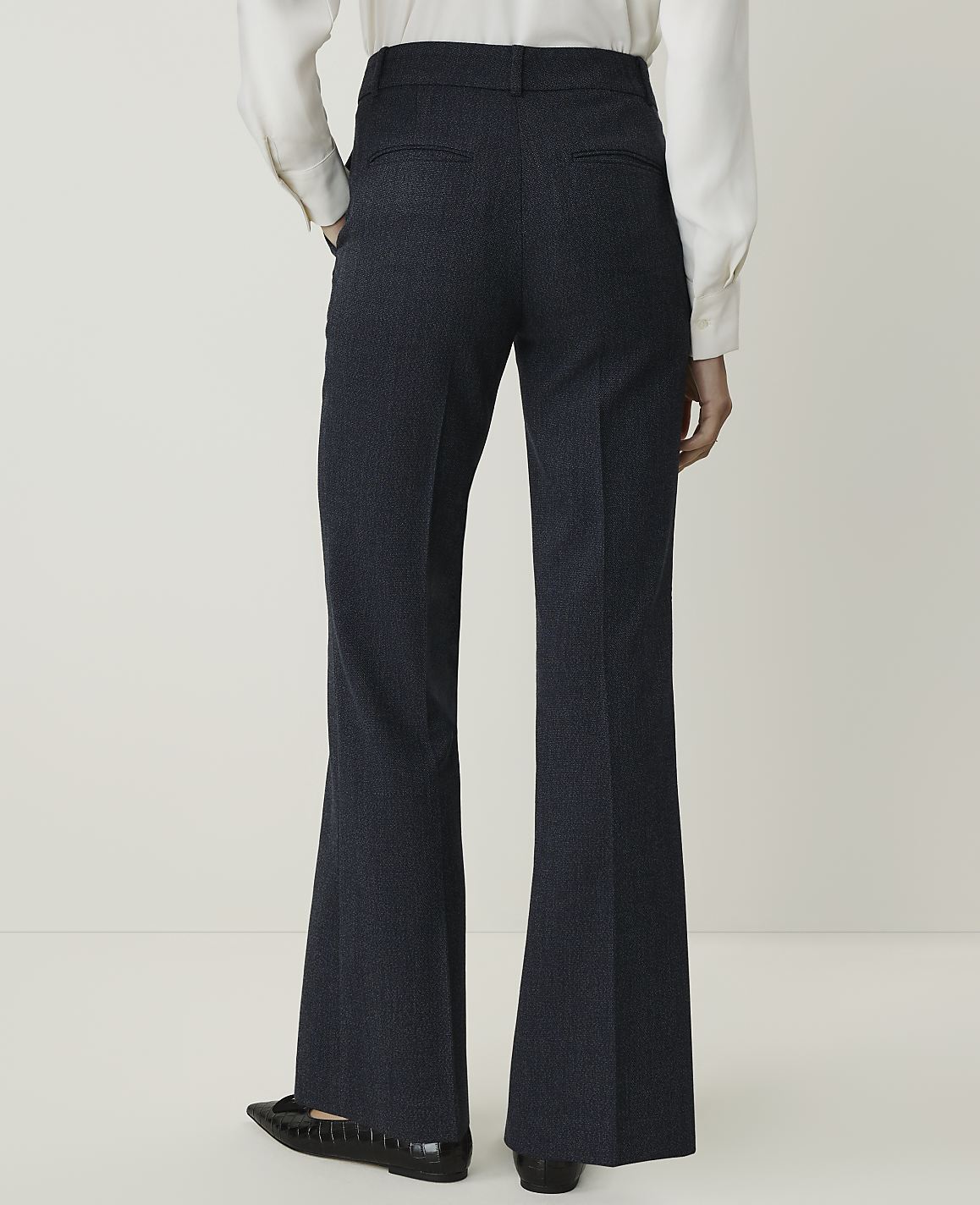 A person wearing dark, flared trousers and a white shirt stands against a plain, light background.