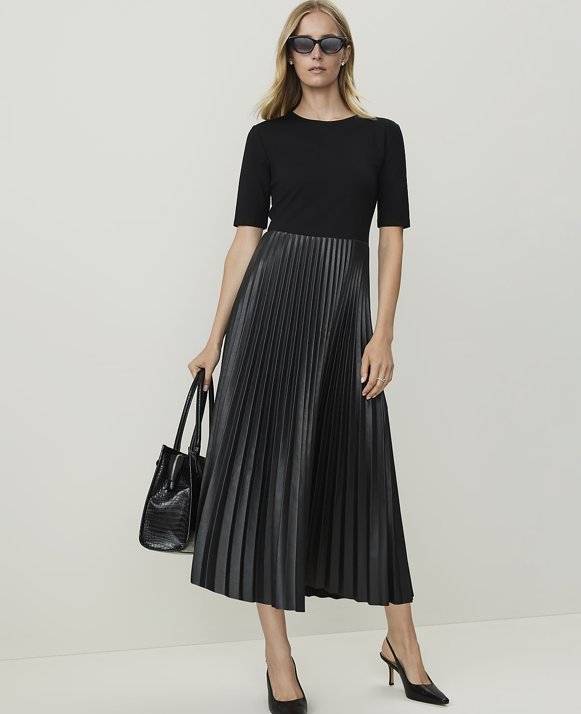 A woman in a black top and pleated faux leather skirt stands against a plain white background, holding a black handbag.