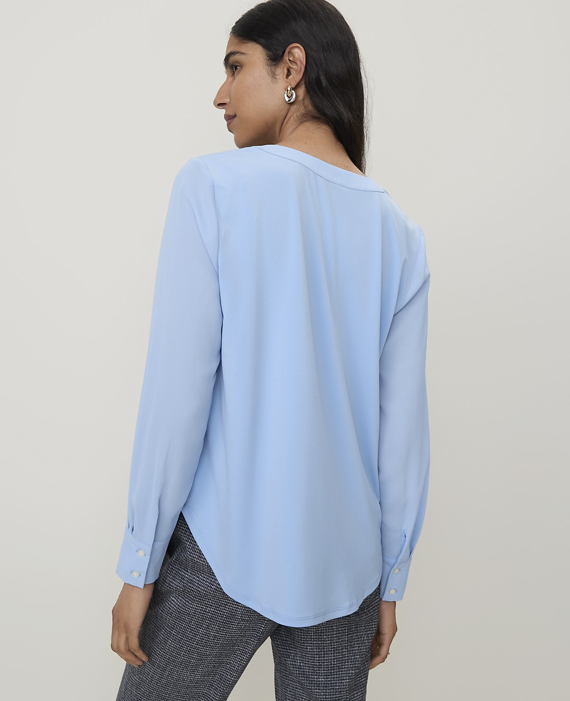 A woman in a light blue blouse and patterned pants stands against a plain, light gray background.
