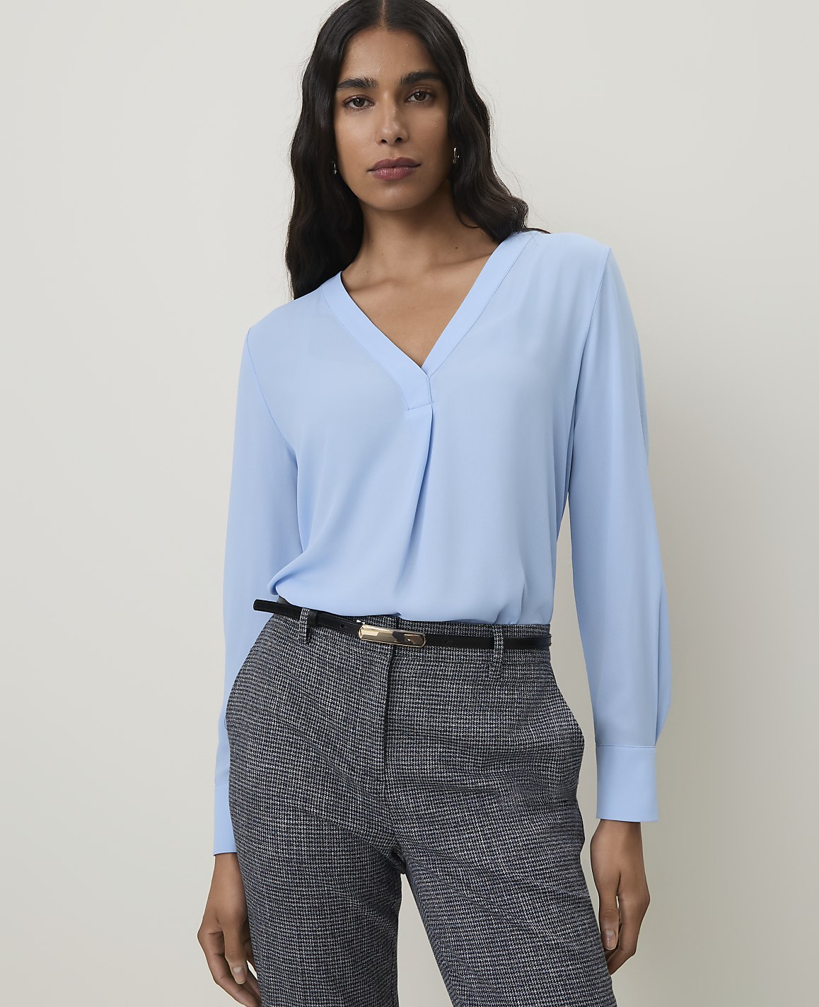 A woman in a light blue blouse and patterned pants stands against a plain background.