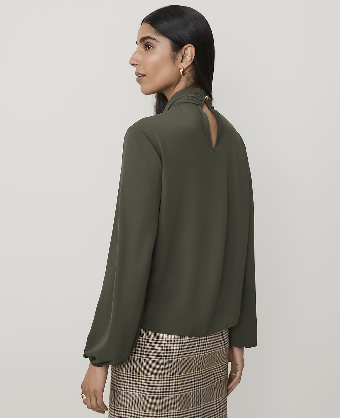 A woman in a green blouse and plaid skirt stands against a plain, light brown background.
